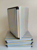 One More Chapter Premium Kindle Case in Rainy Sunday – open folio standing upright on stack of closed cases, dusty blue and cream stripe